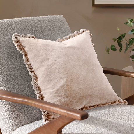 Bertie Velvet Cotton Cushion in natural, shown with a frayed fringed edge on a grey armchair with wooden arms in a living room setting.