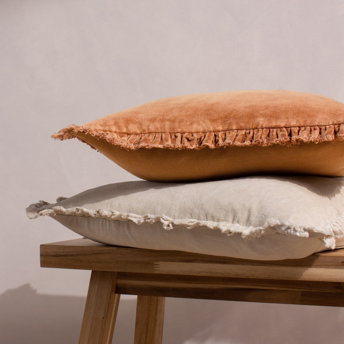 Bertie Velvet Cotton Cushion in natural, shown stacked with a terracotta cushion, featuring a frayed fringed edge on a wooden bench against a plain light background.