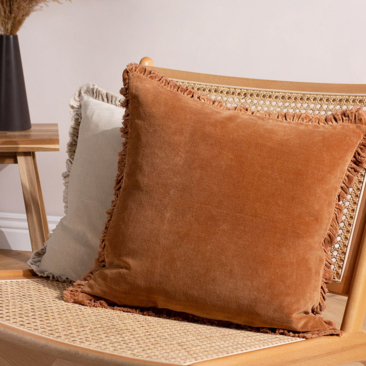 Bertie Velvet Cotton Cushion in rust, a square fringe-edged cushion in warm orange-brown velvet, shown on a rattan chair beside a neutral fringed cushion.