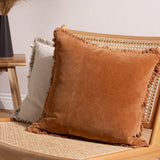 Bertie Velvet Cotton Cushion in rust, a square fringe-edged cushion in warm orange-brown velvet, shown on a rattan chair beside a neutral fringed cushion.