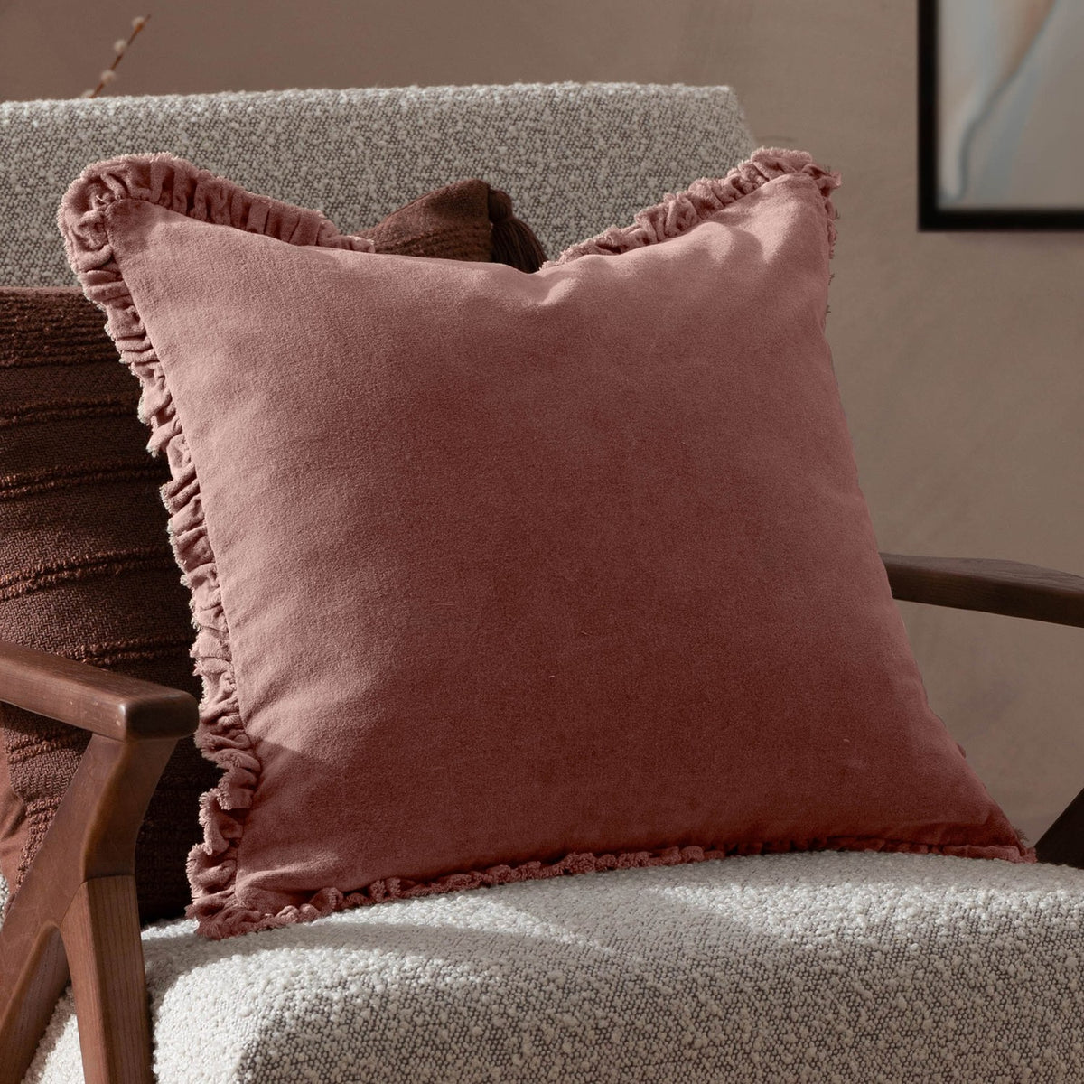 Bertie Velvet Cotton Cushion in rose pink with frayed, ruffled edging, shown on a light bouclé armchair in a living room setting.