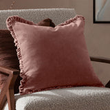 Bertie Velvet Cotton Cushion in rose pink with frayed, ruffled edging, shown on a light bouclé armchair in a living room setting.