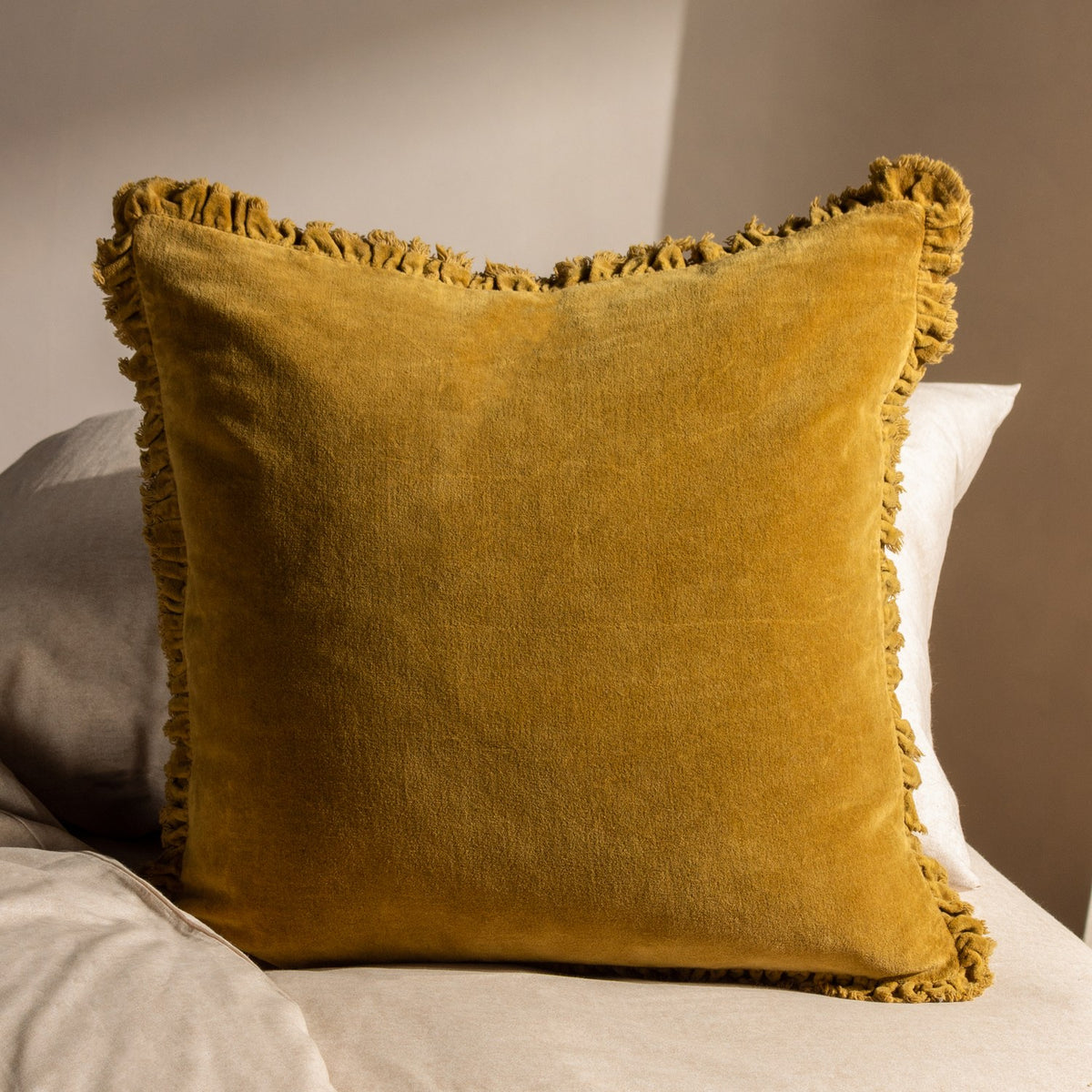Bertie Velvet Cotton Cushion in Saffron, a square 45x45cm cushion with frayed fringed edges, shown propped on a neutral bed setting.
