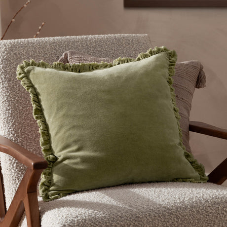Bertie Velvet Cotton Cushion in light sage with ruffled edge trim, shown on a textured armchair in a living room setting.