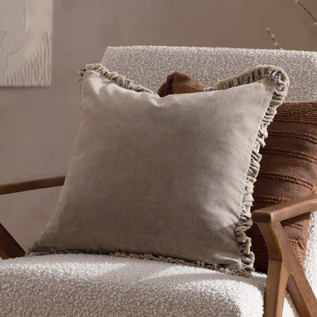 Bertie Velvet Cotton Cushion in pale grey-taupe with a frayed, ruched border edge, shown resting on a boucle armchair beside a rust-toned cushion.