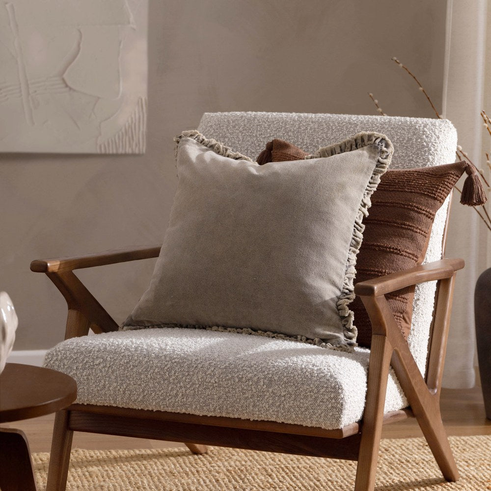 Bertie Velvet Cotton Cushion in pale grey-taupe with ruched, frayed edging, shown on a boucle armchair beside a brown textured cushion in a living room setting.