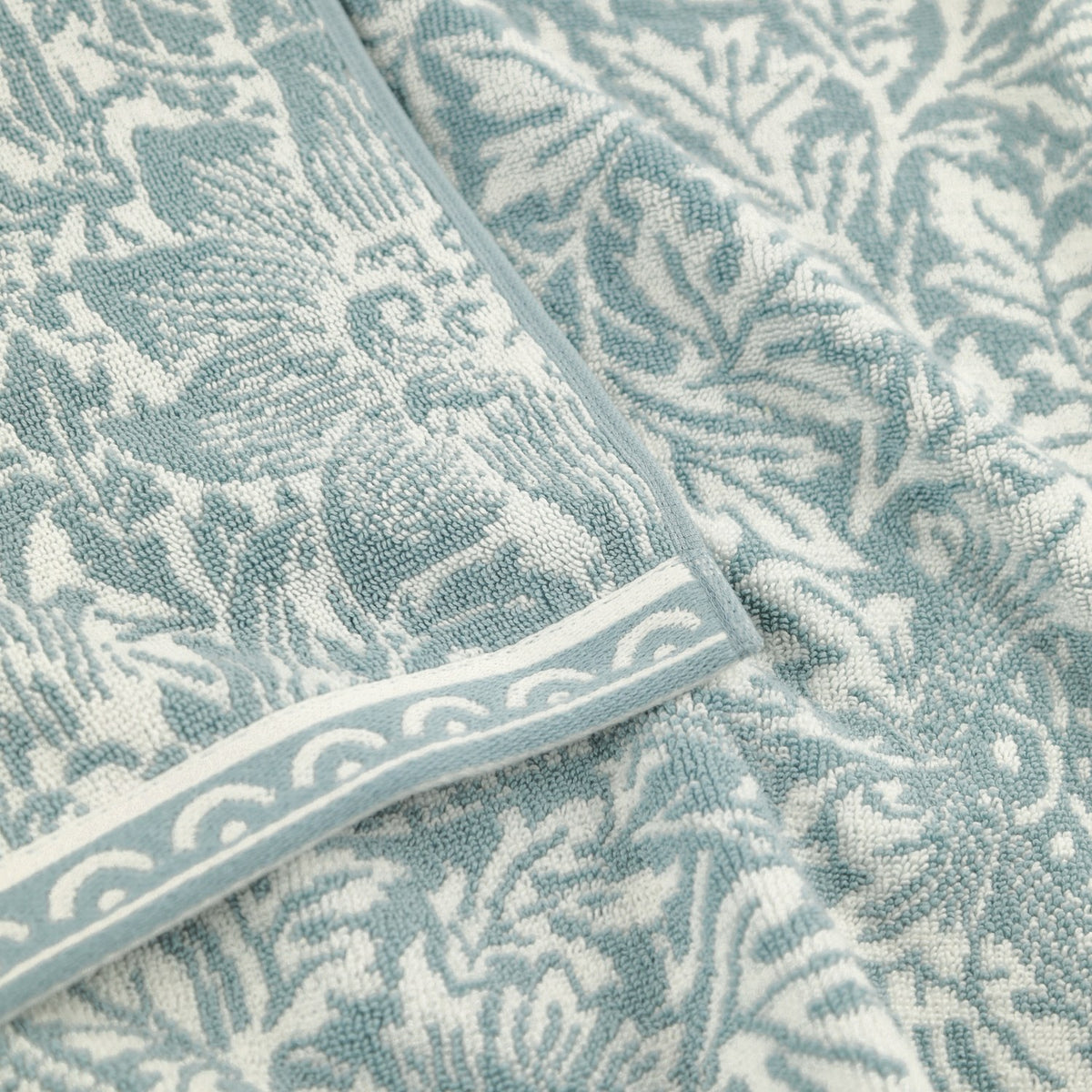Close-up of the Bird & Anemone Jacquard Cotton Towel in slate blue, showing the woven bird and foliage pattern with a scalloped-edge border trim.