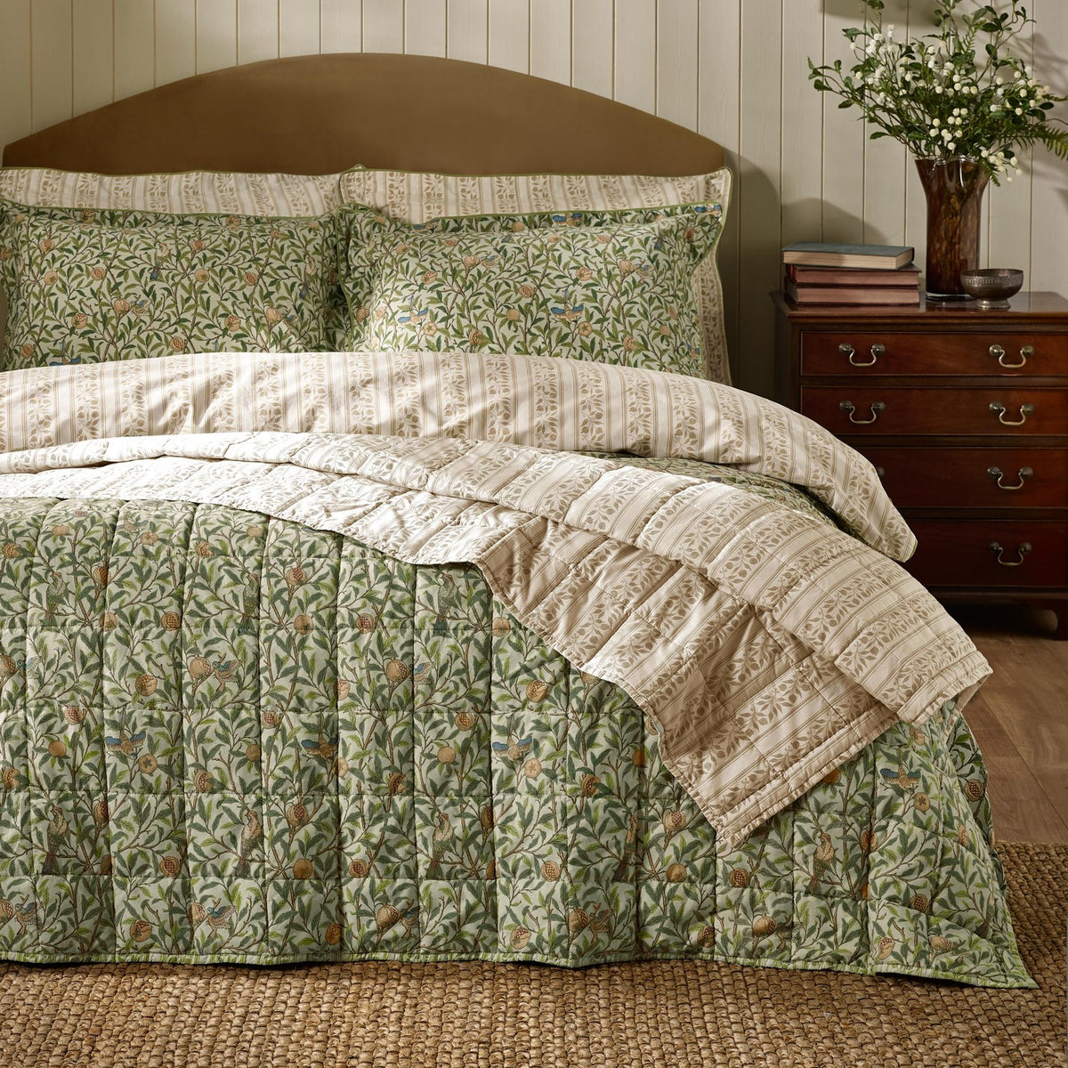 Quilted Bird & Pomegranate cotton bedspread in herbal green with bird and foliage pattern, shown with striped pillowshams on a bed beside a wooden nightstand.