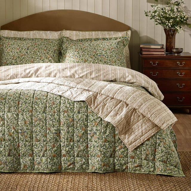 Quilted Bird & Pomegranate cotton bedspread in herbal green with bird and foliage pattern, shown with striped pillowshams on a bed beside a wooden nightstand.