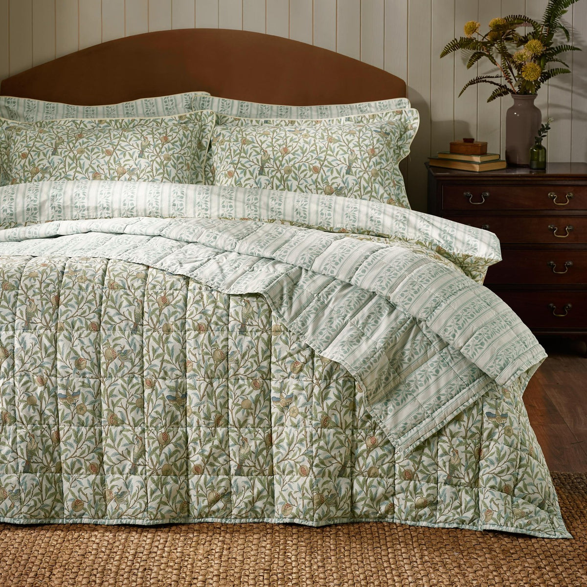Bird & Pomegranate quilted cotton bedspread in sage green with amber and blue floral pattern, shown on a bed with matching pillowcases and striped reverse in a bedroom setting.