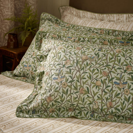 Bird & Pomegranate percale cotton pillowcase pair in Herbal Green, oxford border, styled on a bed with striped bedding, plant and jug in background.