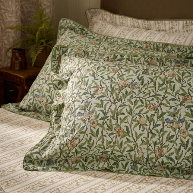 Bird & Pomegranate percale cotton pillowcase pair in Herbal Green, oxford border, styled on a bed with striped bedding, plant and jug in background.
