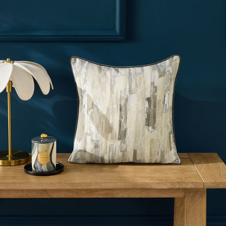 Blocks Velvet Cotton Cushion in silver with abstract vertical block pattern and metallic shimmer, shown on a wooden bench against a dark blue wall.