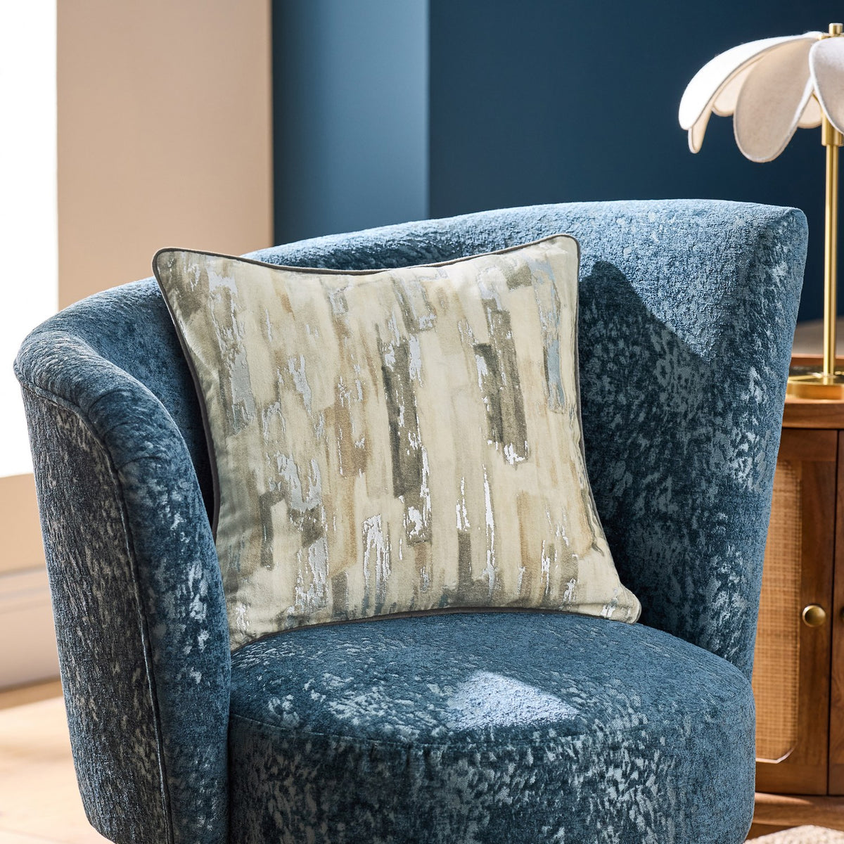Blocks Velvet Cotton Cushion in silver with cream and sandy abstract block pattern, shown on a blue textured armchair in a living room setting.