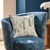 Blocks Velvet Cotton Cushion in silver with cream and sandy abstract block pattern, shown on a blue textured armchair in a living room setting.