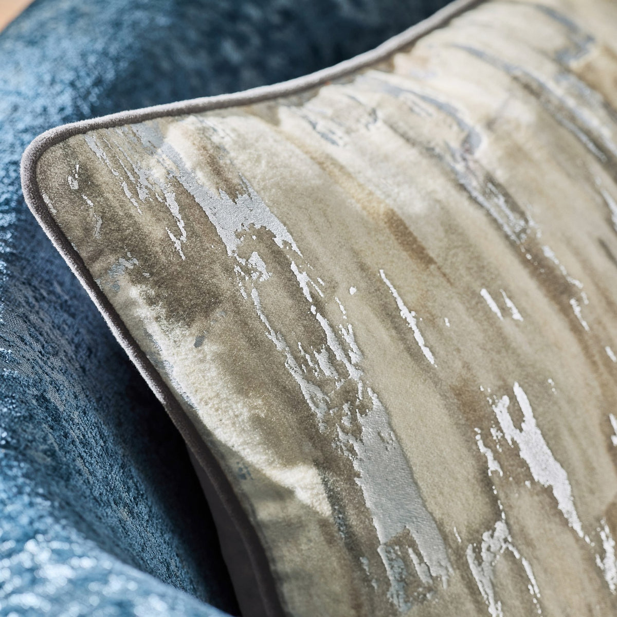 Close-up of the Blocks Velvet Cotton Cushion in silver, featuring a foil-printed abstract vertical stripe design in cream, stone and sandy tones with piped edge, on a blue textured sofa.