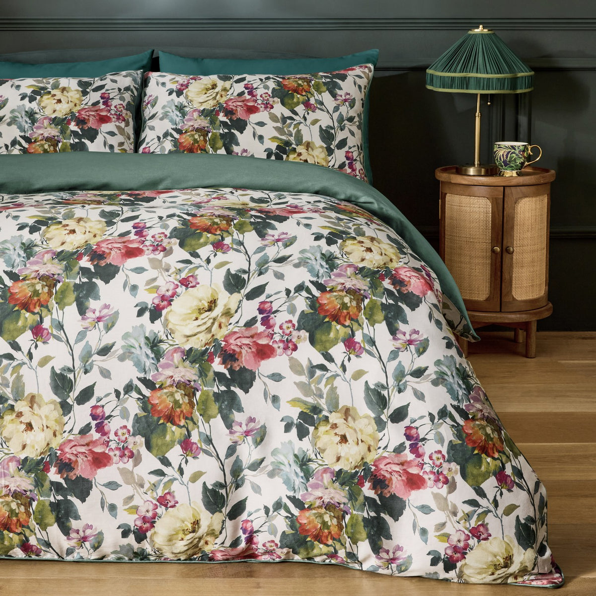 Bloom Cotton Floral Duvet Cover Set in cream with pink, coral and green painterly floral print, shown with matching pillowcases and green reverse on a bed in a dark-walled bedroom.