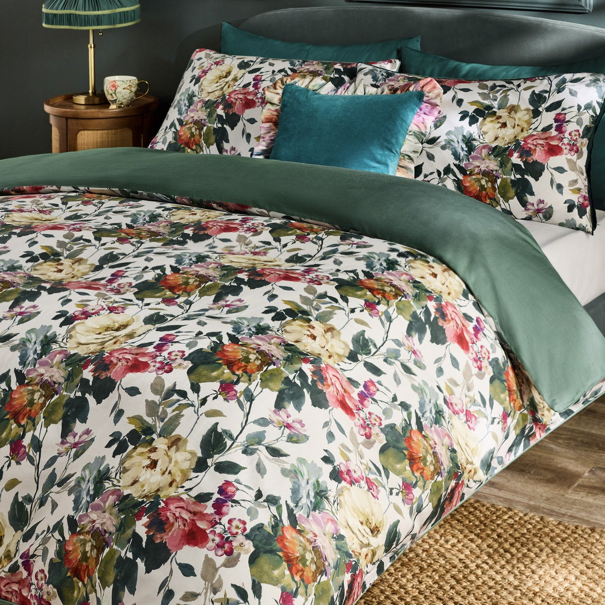 Bloom Cotton Floral Duvet Cover Set in cream with pink, coral and green blooms, styled on a bed with a green reverse, matching pillowcases and a teal velvet cushion.
