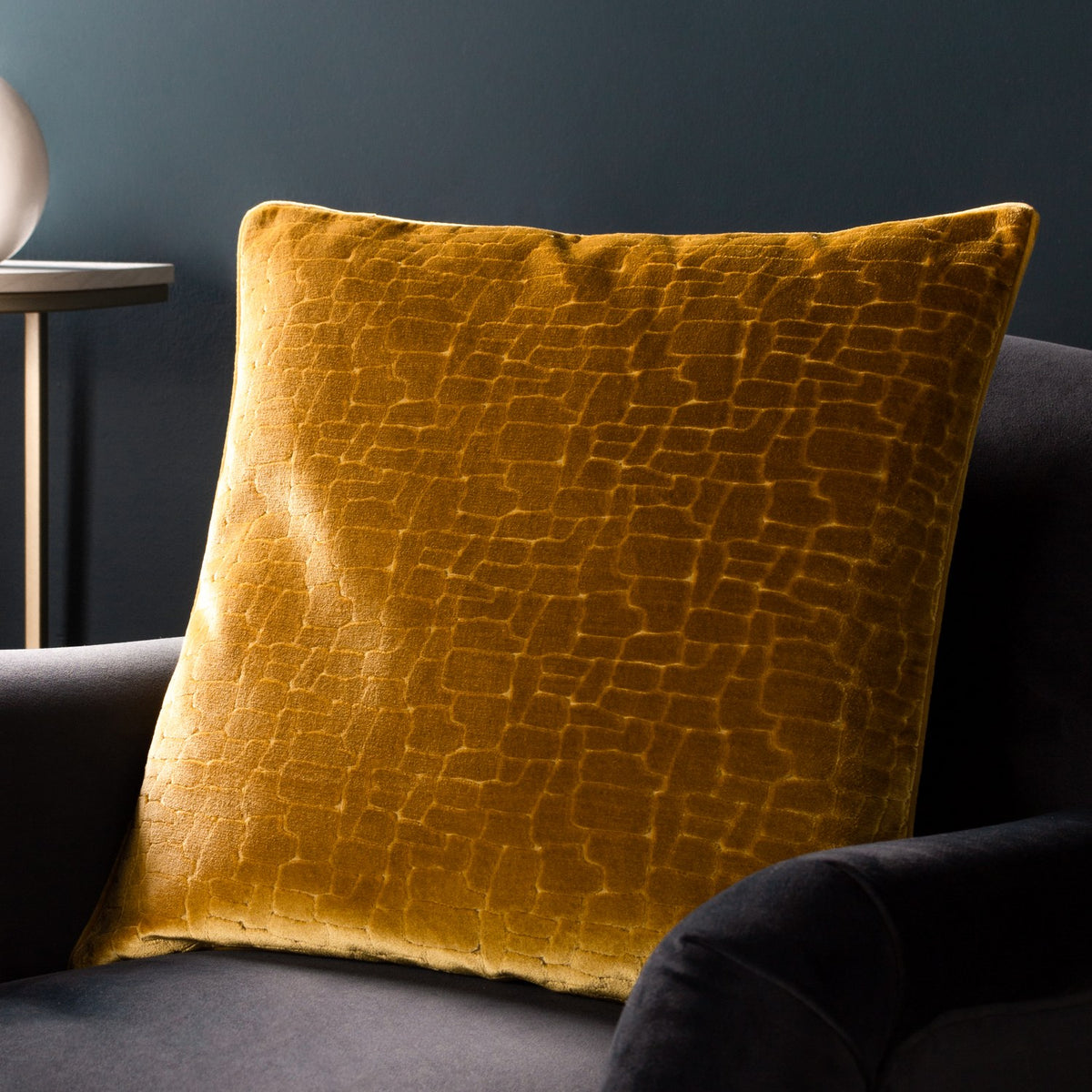 Bloomsbury Abstract Velvet Cushion in mustard, square 50x50cm with raised geometric cut-velvet pattern, styled on a dark grey armchair.