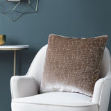 Bloomsbury Abstract Velvet Cushion in taupe with cut-pile cellular pattern, shown on a white armchair against a dark teal wall.