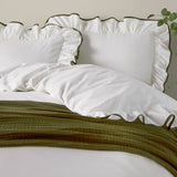 White cotton pillowcases with ruffled edge and olive-green piped trim, styled on a bed with matching duvet and green throw.