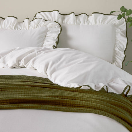 White cotton pillowcases with ruffled edge and olive-green piped trim, styled on a bed with matching duvet and green throw.