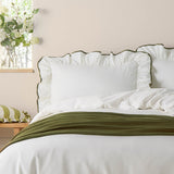 White cotton Blousey pillowcase with ruffled edge and olive-green piped trim, styled on a bed with matching duvet and green throw in a bedroom setting.