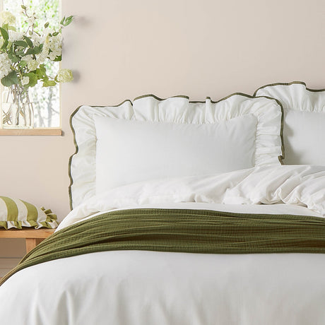 White cotton Blousey pillowcase with ruffled edge and olive-green piped trim, styled on a bed with matching duvet and green throw in a bedroom setting.