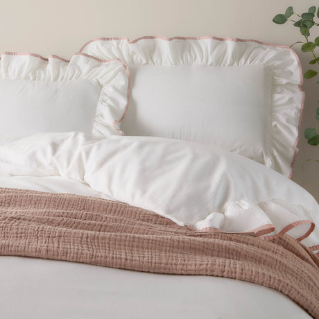 White cotton Blousey pillowcases with ruffled edges and pink piped trim, styled on a bed with a pink throw in a bedroom setting.