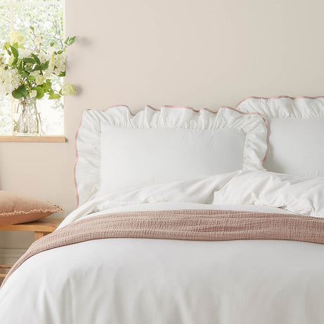 Blousey plain white cotton pillowcase pair with ruffled edge and pink piped trim, shown styled on a bed in a bedroom setting with a pink cotton throw.
