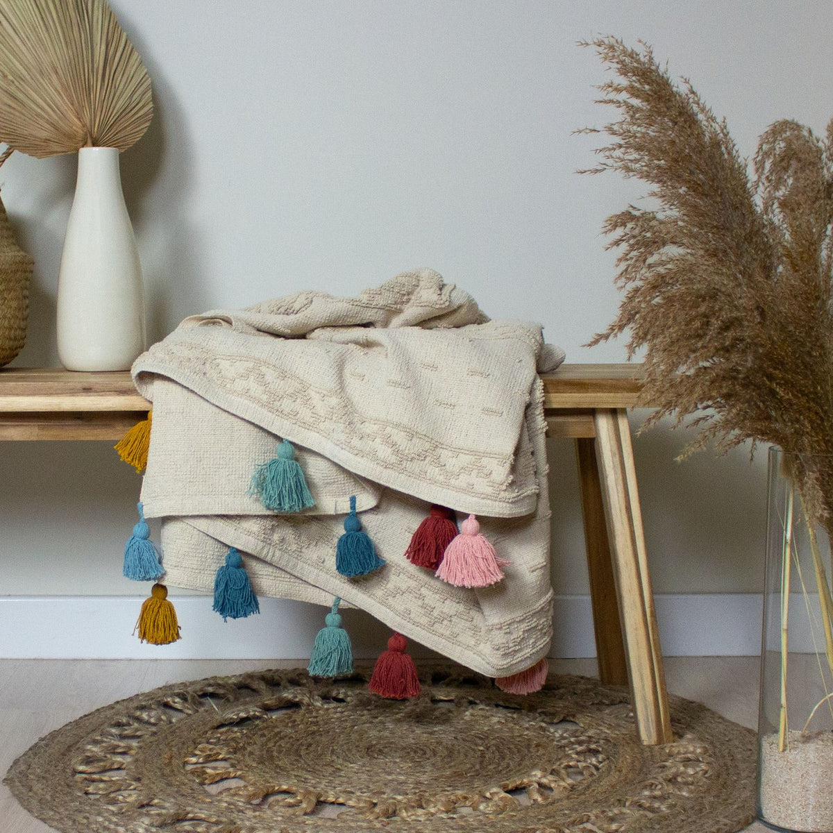 Boho Geometric Cotton Throw in natural cream with tufted geometric bands, draped over a wooden bench with mustard, teal, pink and red tassels along the edge.