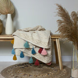 Boho Geometric Cotton Throw in natural cream with tufted geometric bands, draped over a wooden bench with mustard, teal, pink and red tassels along the edge.
