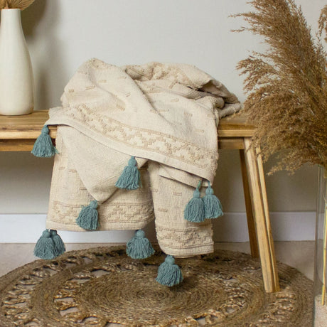 Boho Geometric Cotton Throw in cream with tufted patterned border and seafoam tassel trim, draped over a wooden bench beside pampas grass.