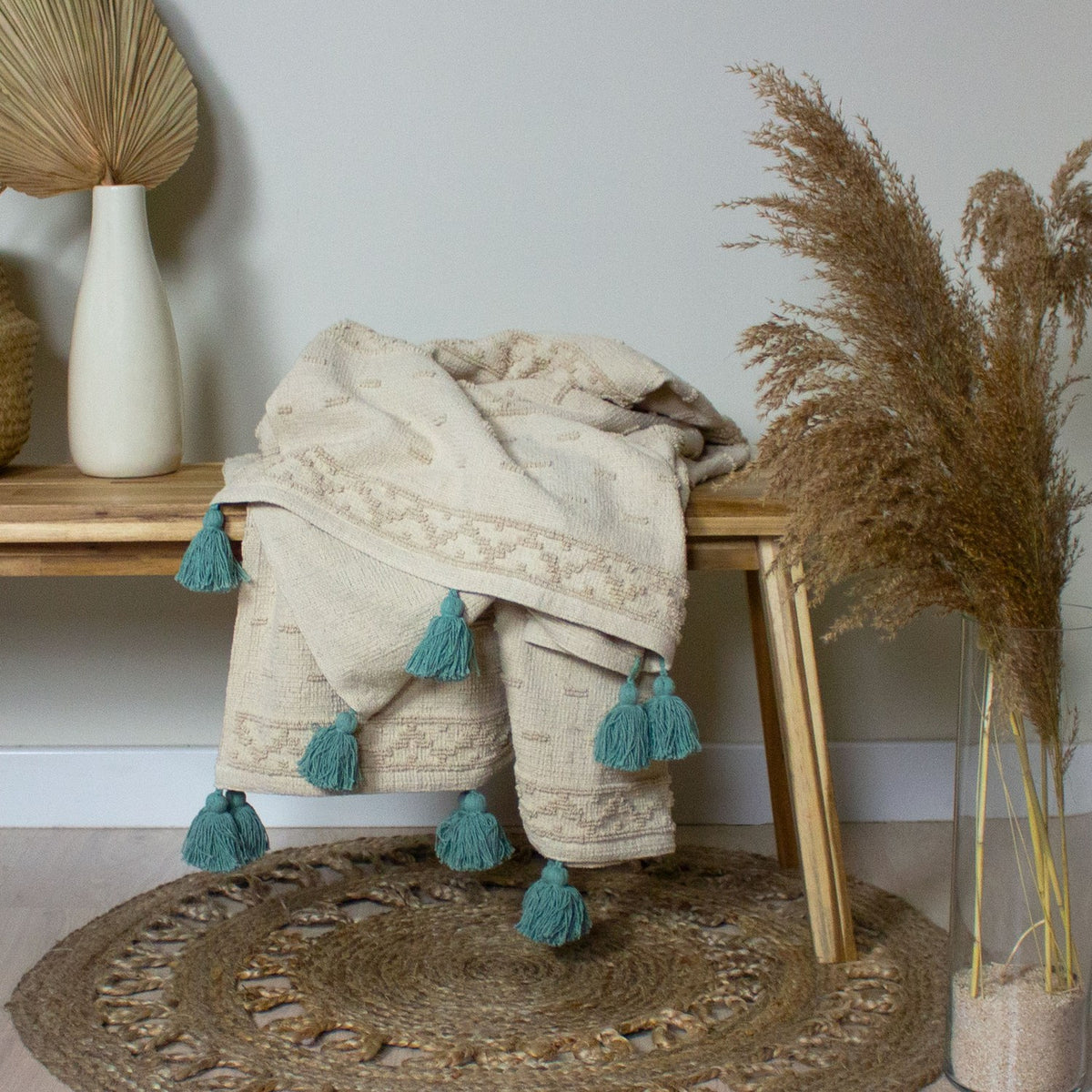 Boho Geometric Cotton Throw in Seafoam, cream tufted textured fabric with teal tassel trim, draped over a wooden bench beside dried pampas grass.