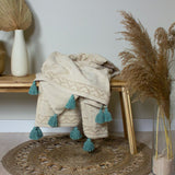 Boho Geometric Cotton Throw in Seafoam, cream tufted textured fabric with teal tassel trim, draped over a wooden bench beside dried pampas grass.