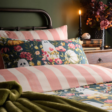 Bootanical reversible duvet cover set on a bed, showing pink and cream striped side and reverse with teal ghost and floral pumpkin print, styled with a green throw and candlelit bedside display.