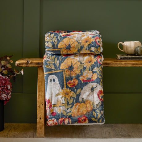 Botanical floral polyester throw in multi-color autumnal ghost print with teal, gold, rust, and cream, sherpa fleece reverse, draped over a wooden bench against a green panelled wall.