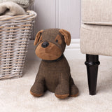 Boston Faux Fur Cotton Door Stop in Brown, shaped as a seated dog with herringbone woven body, faux fur face and paws, and faux leather snout and collar, shown beside a wicker basket and sofa leg.