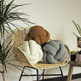 Boucle Knot Plain Polyester Cushion in Ecru, shown in a woven rattan chair alongside brown and grey knot cushions, in a room with plants.