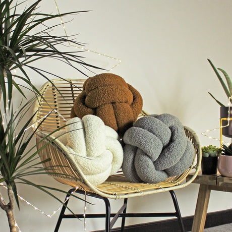 Boucle Knot Plain Polyester Cushion in Ecru, shown in a woven rattan chair alongside brown and grey knot cushions, in a room with plants.