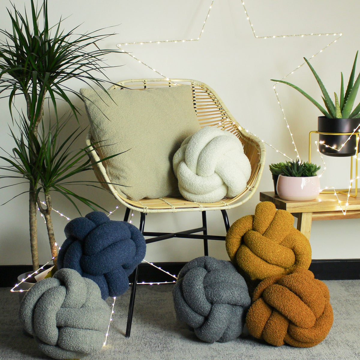 Ecru Boucle Knot cushion on a rattan chair, styled with other knot cushions in navy, grey, and mustard, plus a plain cream cushion, in a room with plants and fairy lights.