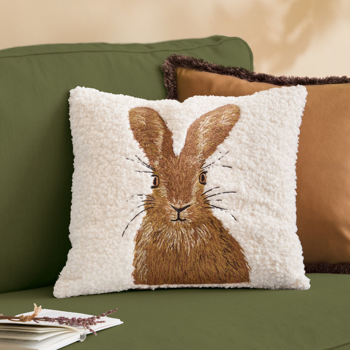 Square natural boucle cushion with embroidered brown hare design, shown on a green sofa alongside tan and brown accent cushions.