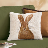 Square natural boucle cushion with embroidered brown hare design, shown on a green sofa alongside tan and brown accent cushions.