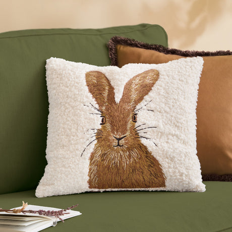 Square natural boucle cushion with embroidered brown hare design, shown on a green sofa alongside tan and brown accent cushions.