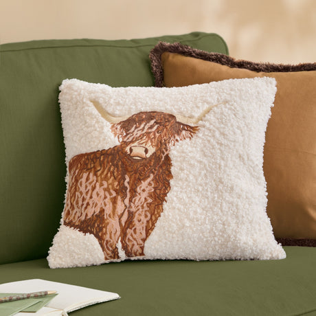 Square boucle cushion with an embroidered Highland cow in brown and tan tones on a cream textured base, shown on a green sofa beside a tan cushion.