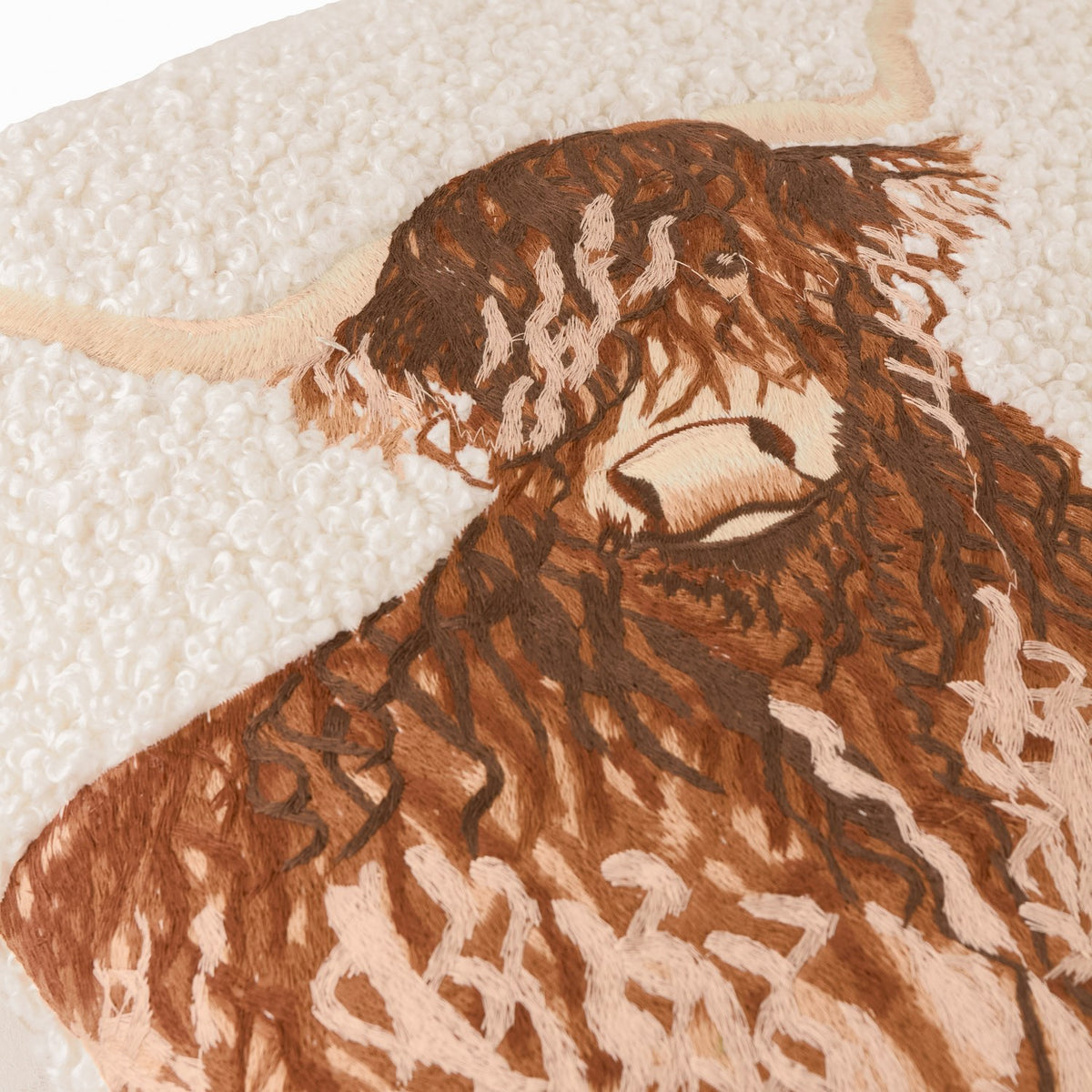 Close-up of the Boucle Highland Cow cushion showing embroidered brown and tan fringe detail with pale horns on a cream textured boucle base.