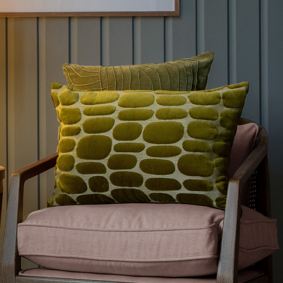 Boulder Geometric Velvet Cushion in olive with raised pebble-shaped pattern, styled on an armchair with a striped olive velvet cushion behind it.