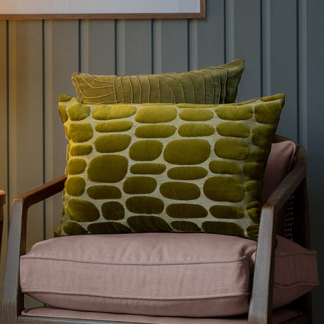 Boulder Geometric Velvet Cushion in olive with raised pebble-shaped pattern, styled on an armchair with a striped olive velvet cushion behind it.