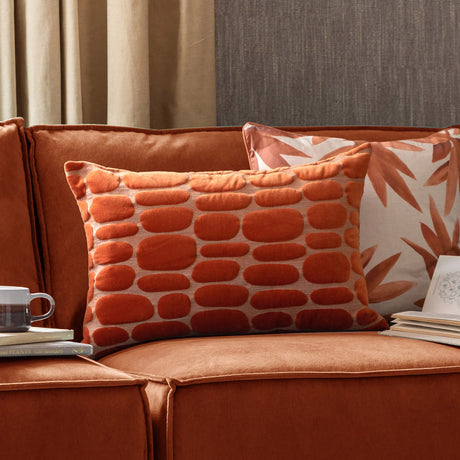 Boulder Geometric Velvet Cushion in Sunset orange with raised pebble-like pattern, shown on a rust velvet sofa alongside a leaf-print pillow.