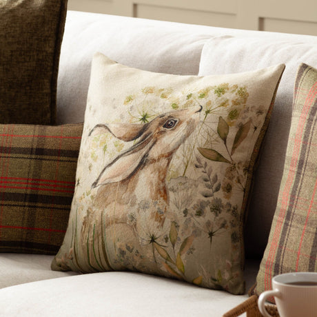 Bramble Hare square cushion in beige and green polyester fabric with a printed hare among watercolour cow parsley, styled on a sofa with tartan cushions.